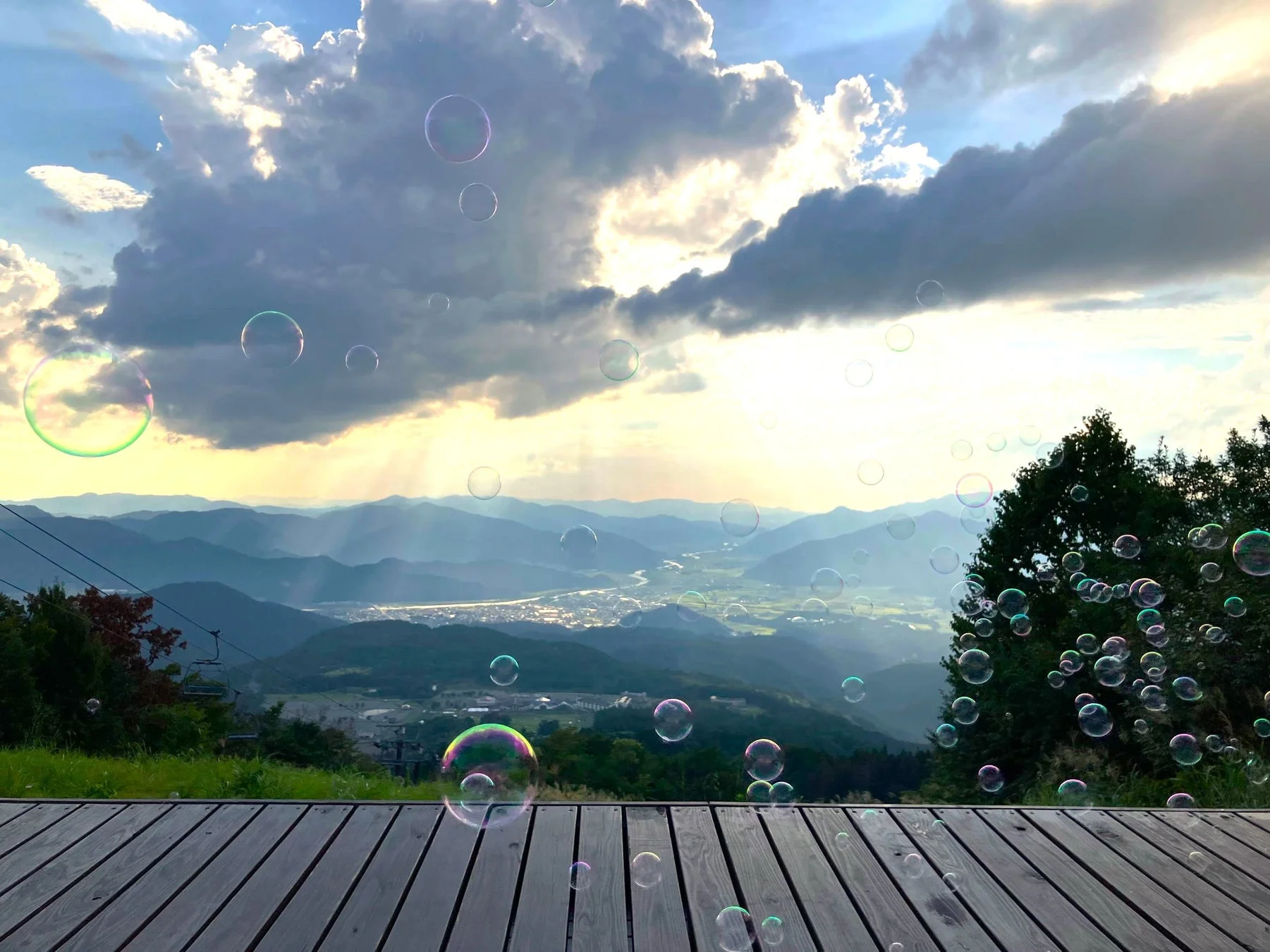 標高の高い場所から見下ろす美しい山々の風景。空には厚い雲と太陽光が差し込み、景色全体にシャボン玉が舞い、幻想的な雰囲気を醸し出しています。