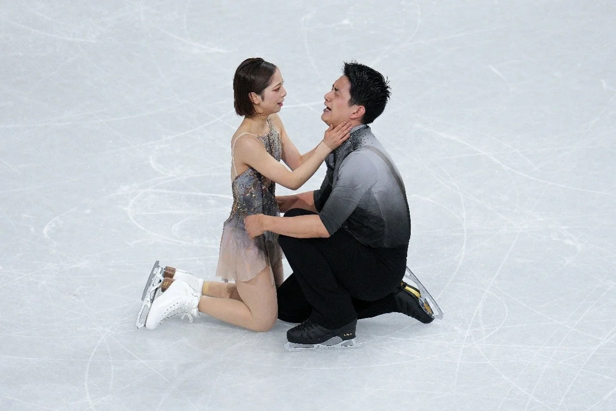 フィギュアスケートのペア選手が氷上で膝をつき、女性が男性の顔に手を添え、お互いの感情を分かち合っている様子。