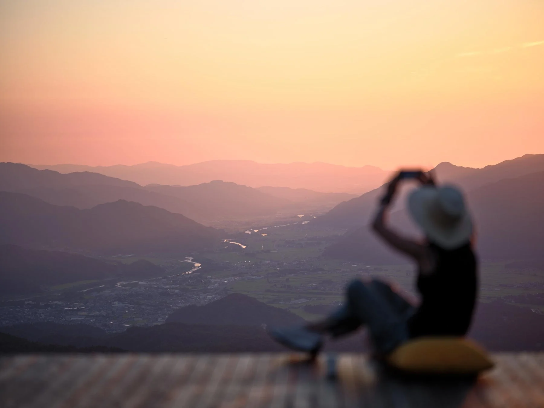 夕焼けに染まる山並みと谷間を望む絶景を、座ってスマートフォンで撮影している人物の姿。川が流れる町並みが広がる雄大な風景が印象的です。