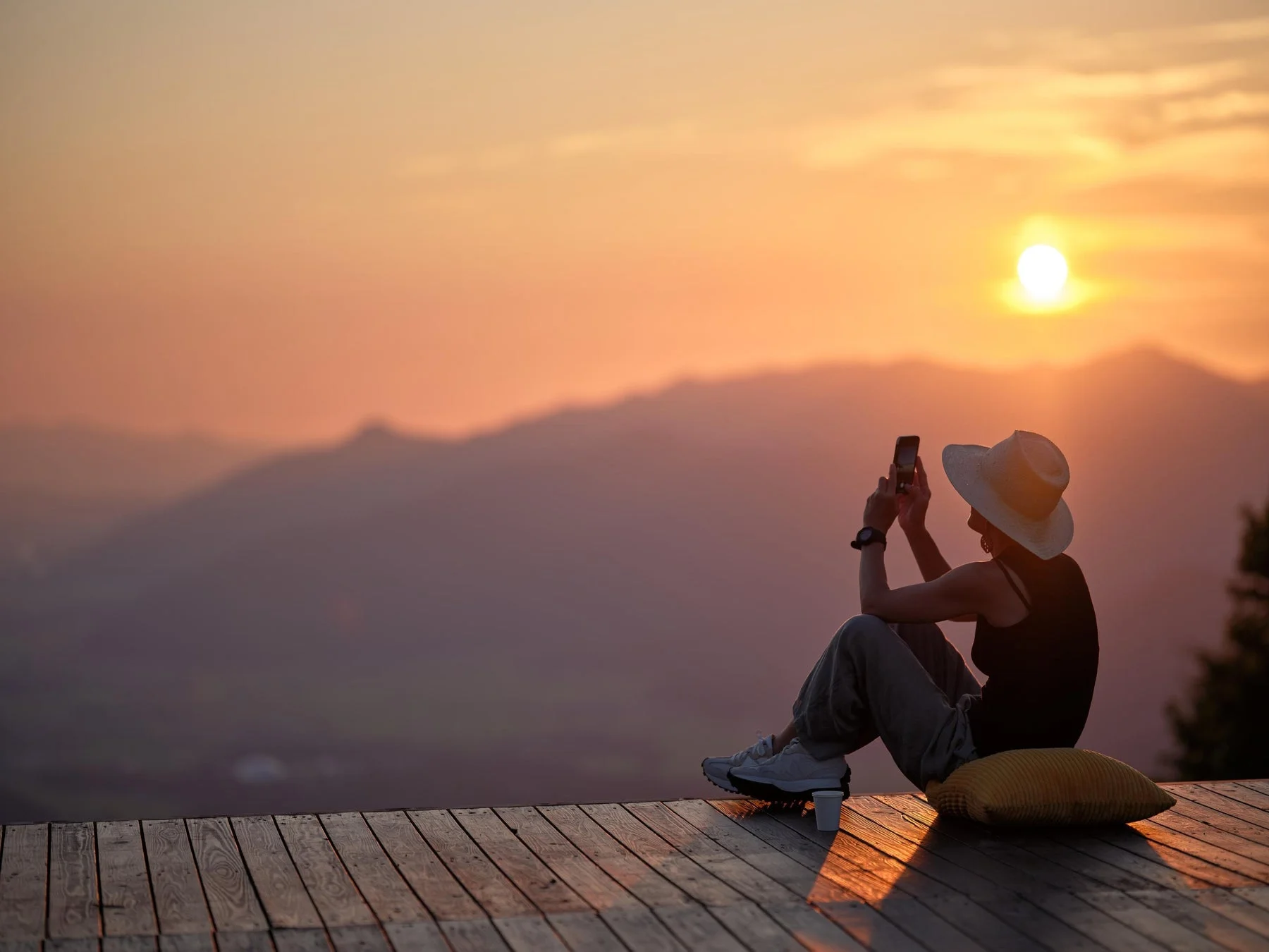 木製デッキに座った女性が、夕焼け空と遠くの山々を背景にスマートフォンで写真を撮っています。