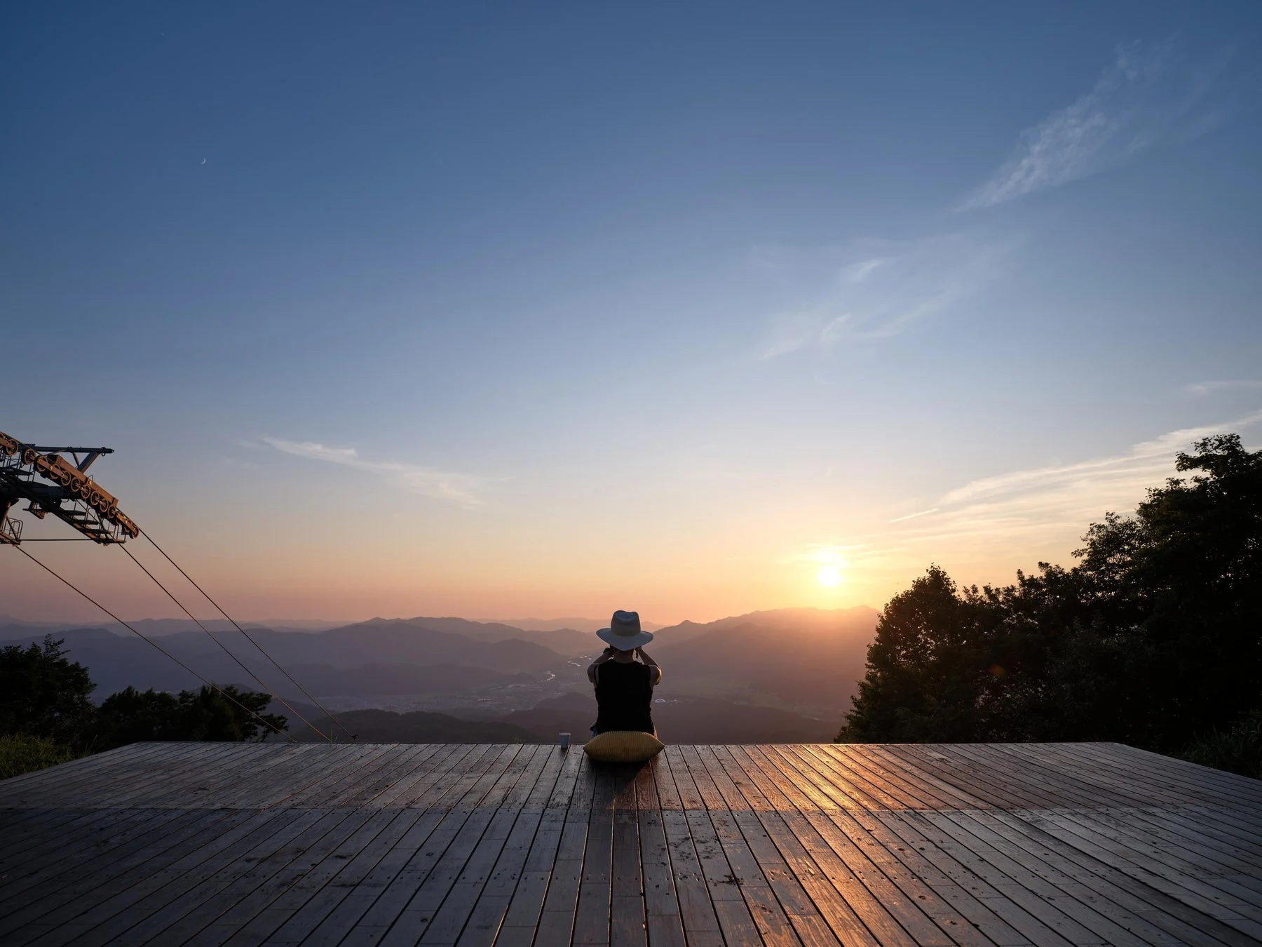 山々の絶景が広がる展望デッキで、夕日または朝日を眺める人物の後ろ姿。